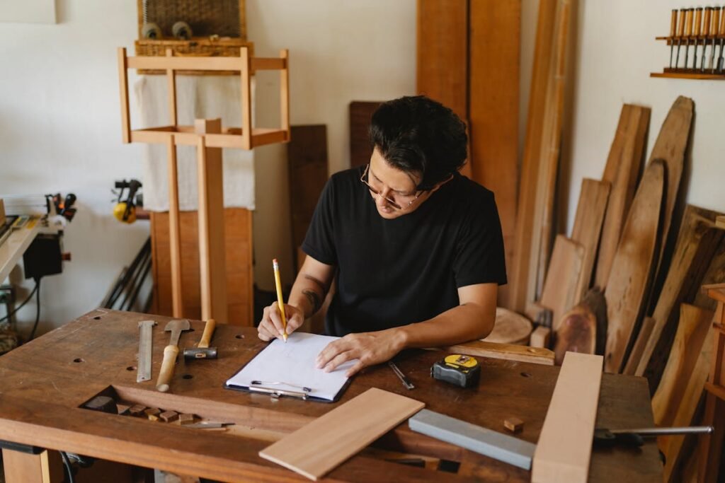 A person with 25 years of experience drafting plans on paper at a woodworking bench surrounded by tools and wood pieces.