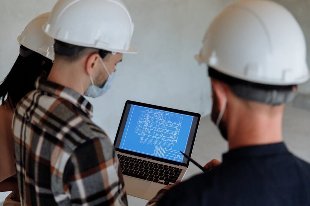 Two construction workers with hard hats from Midtown Builders Group, known for creating exceptional homes, examining blueprints on a laptop.